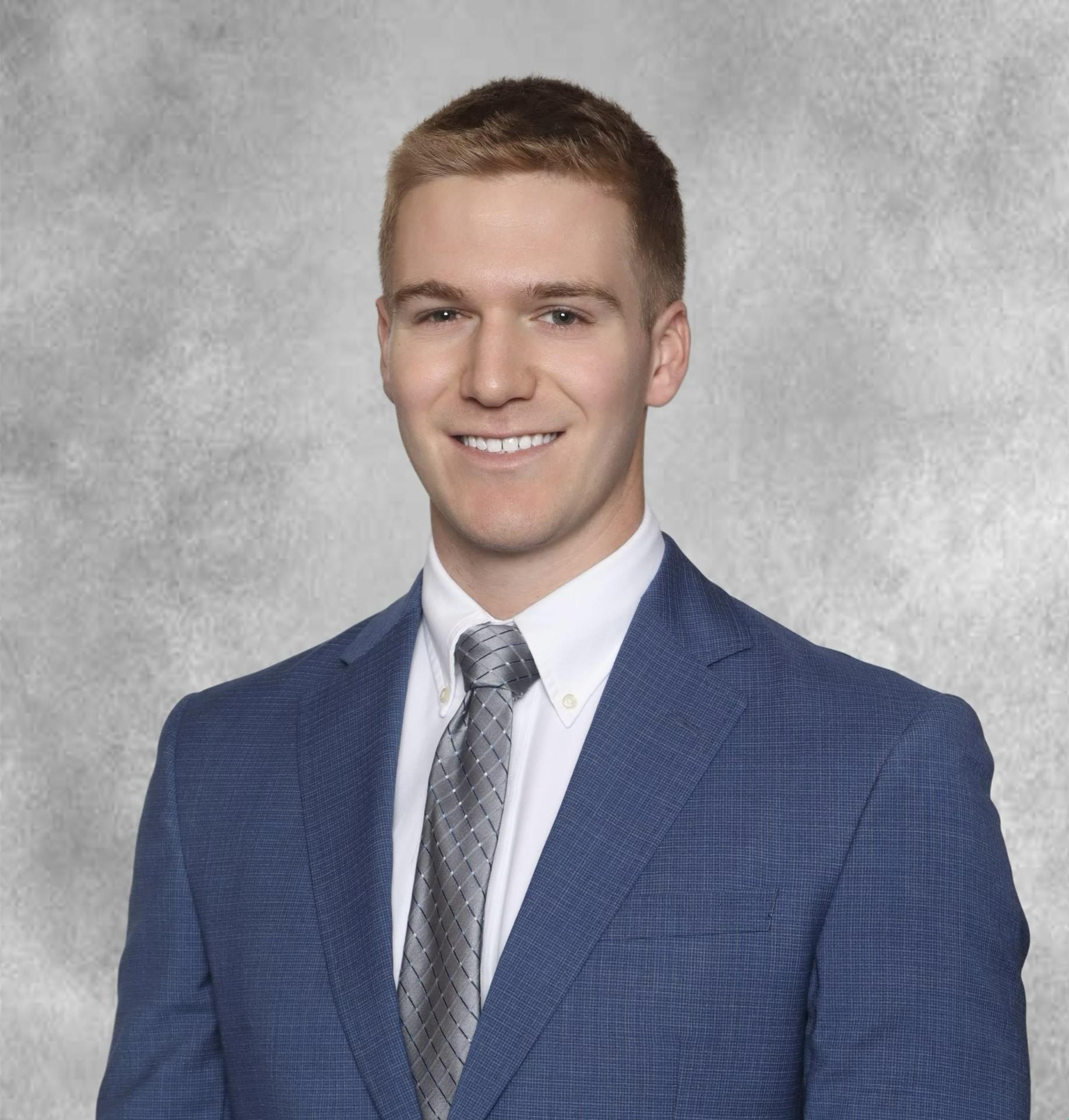 A head-and-shoulders studio portrait of a person wearing a blue suit jacket, a white dress shirt, and a gray patterned tie. The background is a softly textured light gray studio backdrop, and the subject is facing forward in a formal, professional pose.