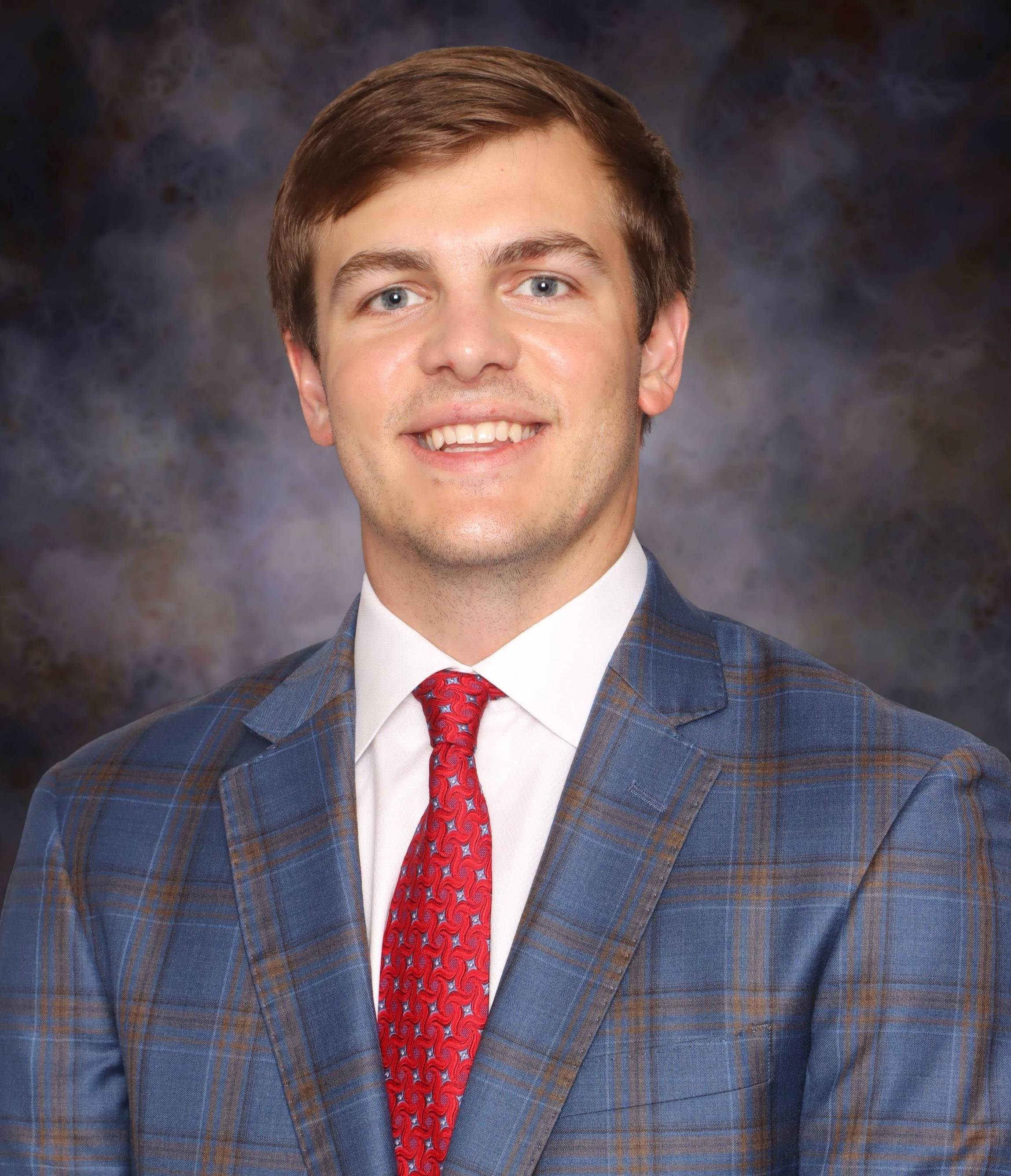A head-and-shoulders studio portrait of a person wearing a blue plaid suit jacket, a white dress shirt, and a red patterned tie. The background is a softly blended dark studio backdrop with gray and brown tones, and the subject is facing forward in a formal, professional pose.