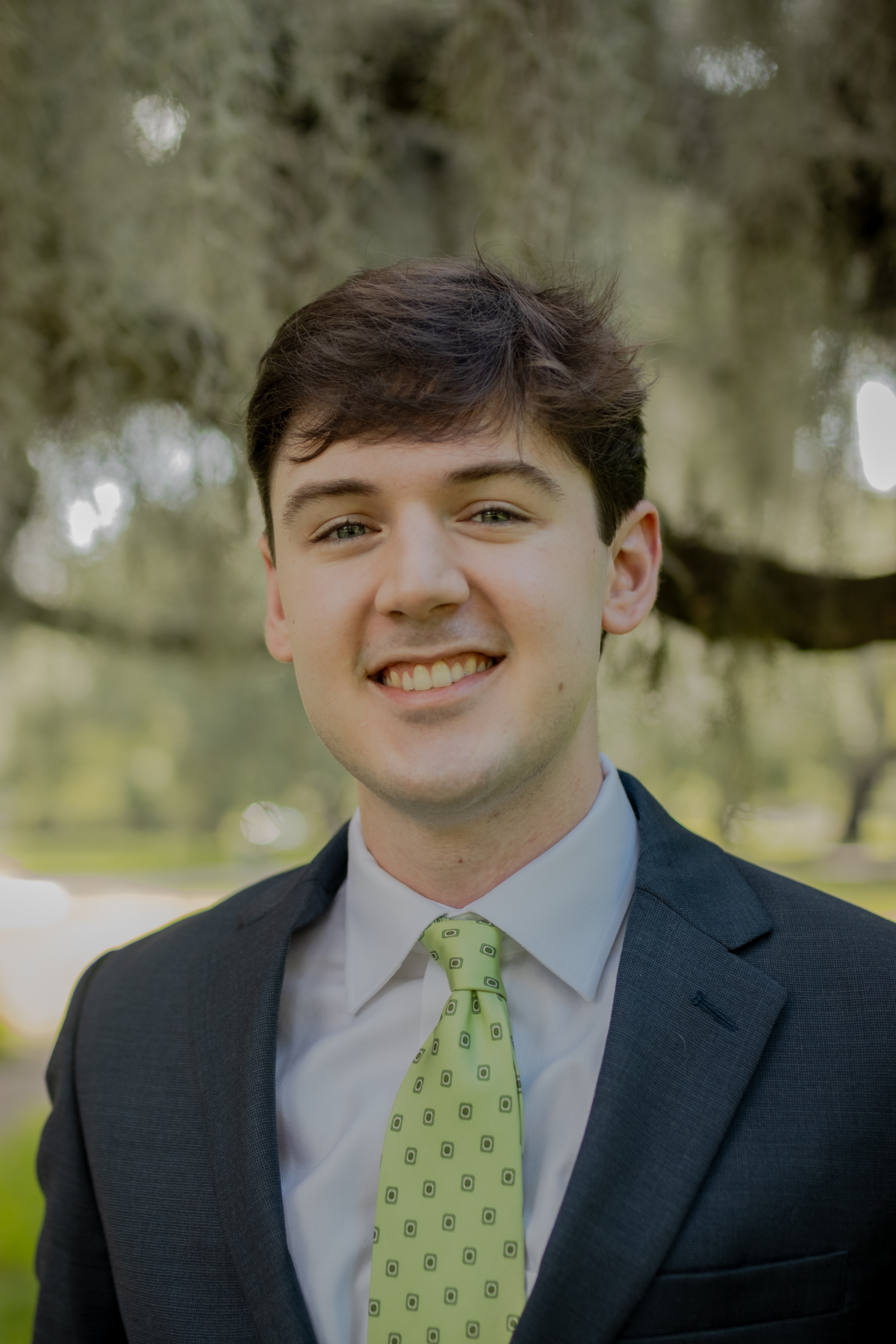 Head-and-shoulders outdoor portrait of a person wearing a dark suit jacket, light dress shirt, and light green patterned tie, facing the camera. Trees with hanging foliage are visible in the softly blurred background.