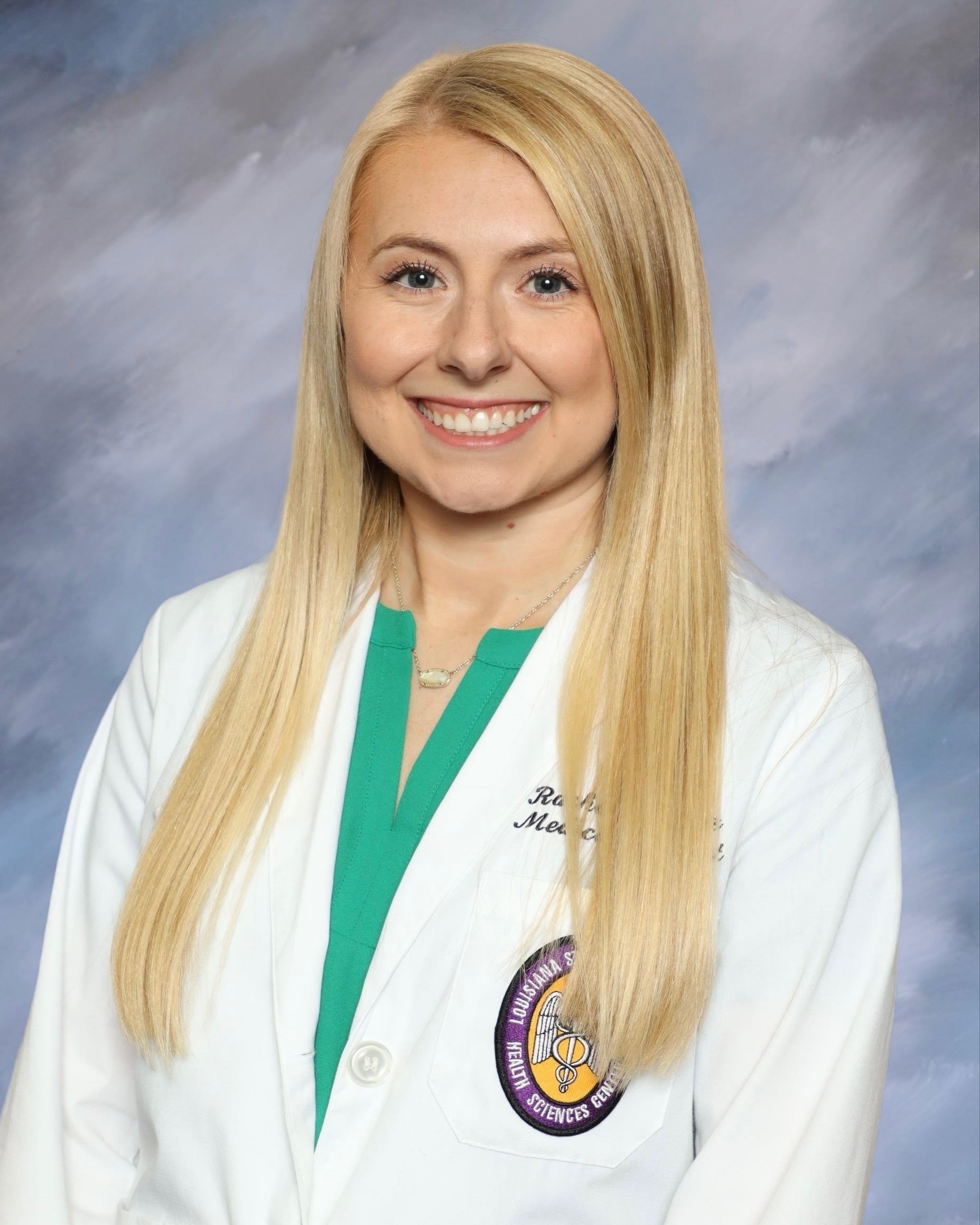 Head-and-shoulders studio portrait of a person with long straight hair wearing a white medical-style coat over a green top, facing the camera against a blue-gray mottled background. A round institutional emblem is visible on the coat.
