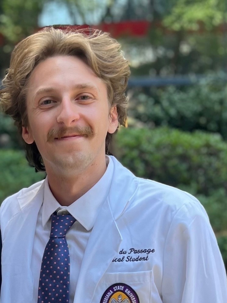 Head-and-shoulders outdoor portrait of a person wearing a white medical-style coat over a dress shirt and patterned tie, facing the camera. Green shrubs and trees appear softly blurred in the background, and an institutional emblem and embroidered text are visible on the coat.