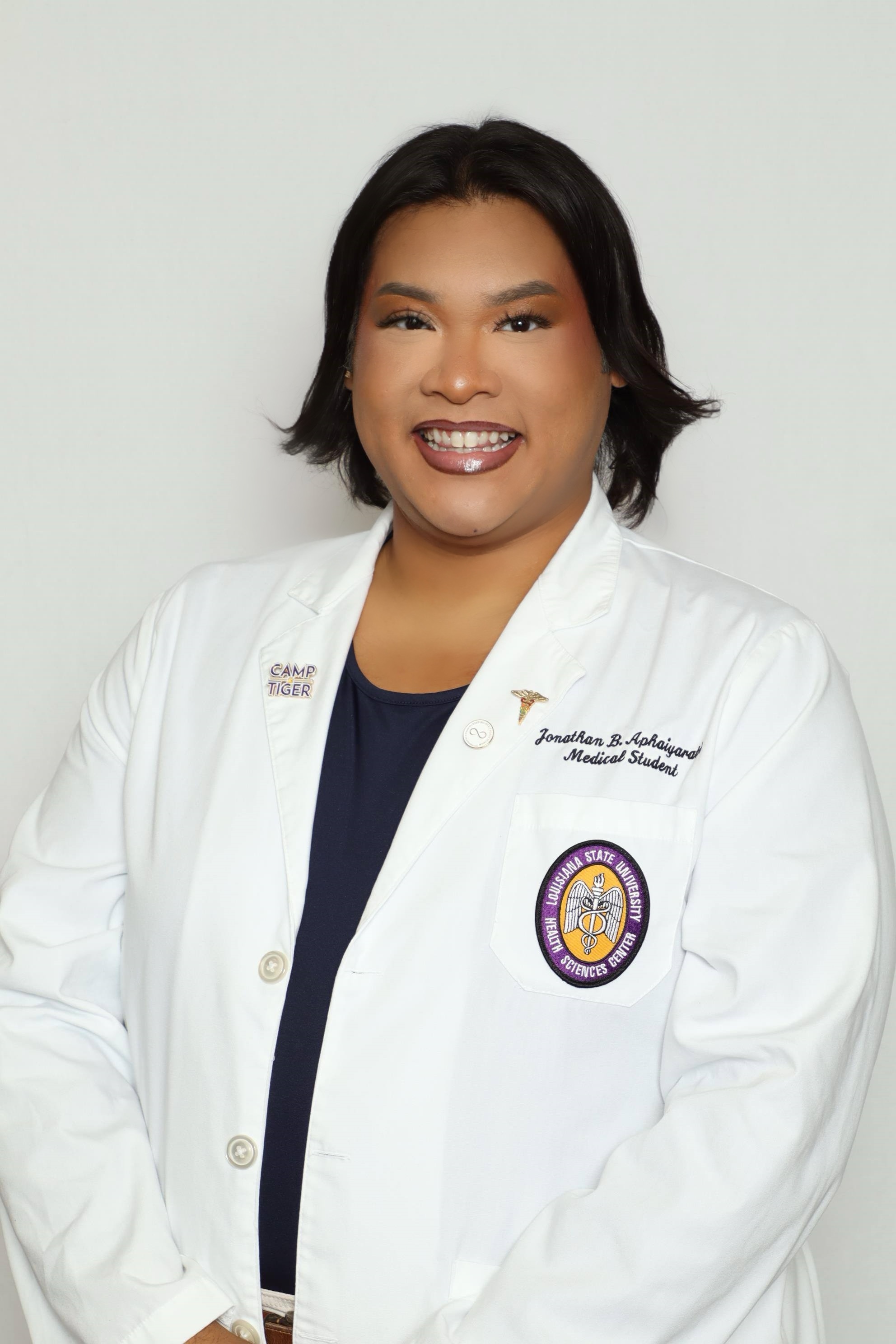 Head-and-shoulders studio portrait of a person with short dark hair wearing a white medical-style coat over a dark top, standing against a plain light background. An institutional emblem and small embroidered text are visible on the coat.