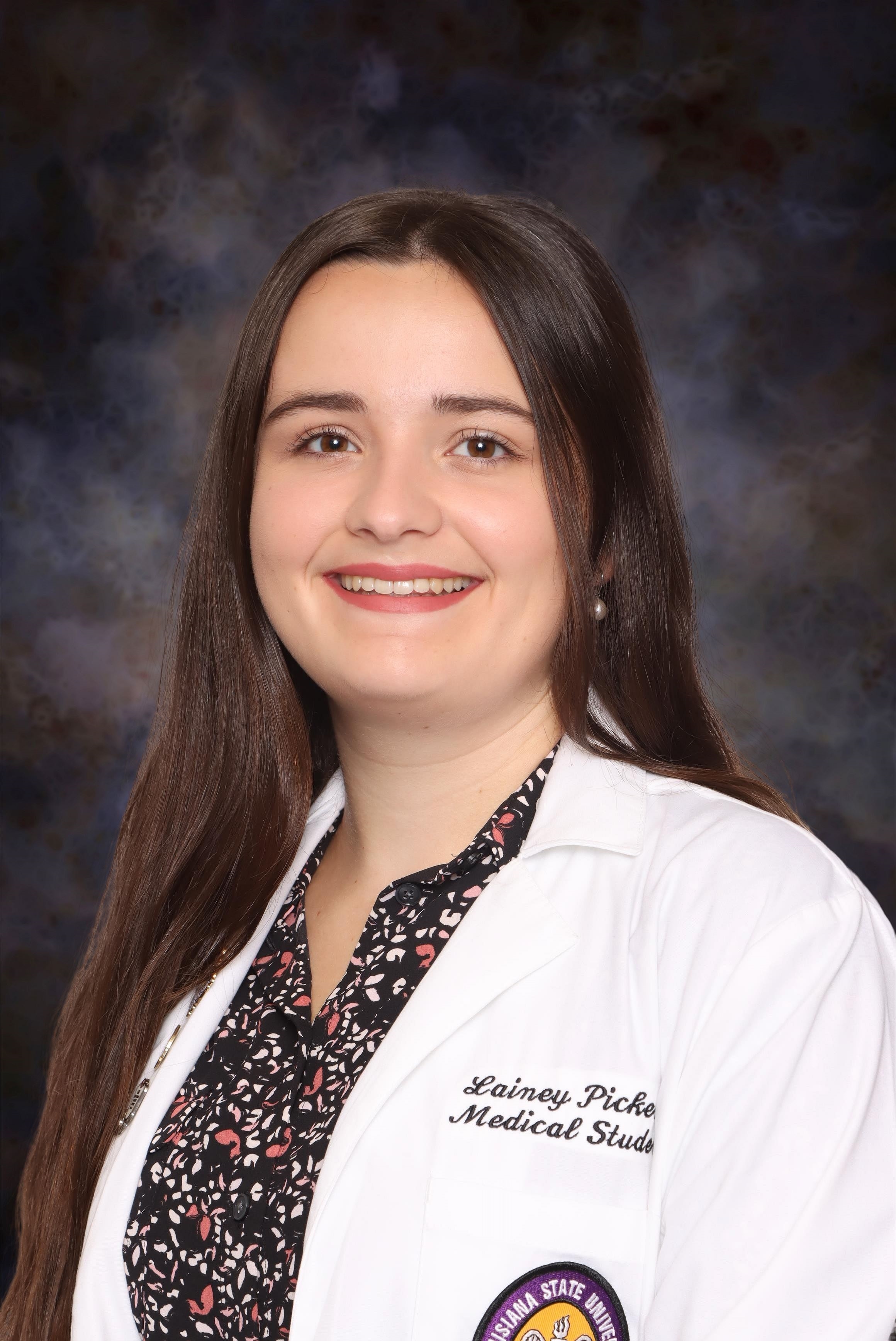 A head-and-shoulders studio portrait of a person wearing a white medical coat over a dark patterned blouse. The medical coat has embroidered text and a circular patch on the sleeve. The background is a softly blurred gray and blue mottled backdrop, and the subject is facing forward in a professional pose.