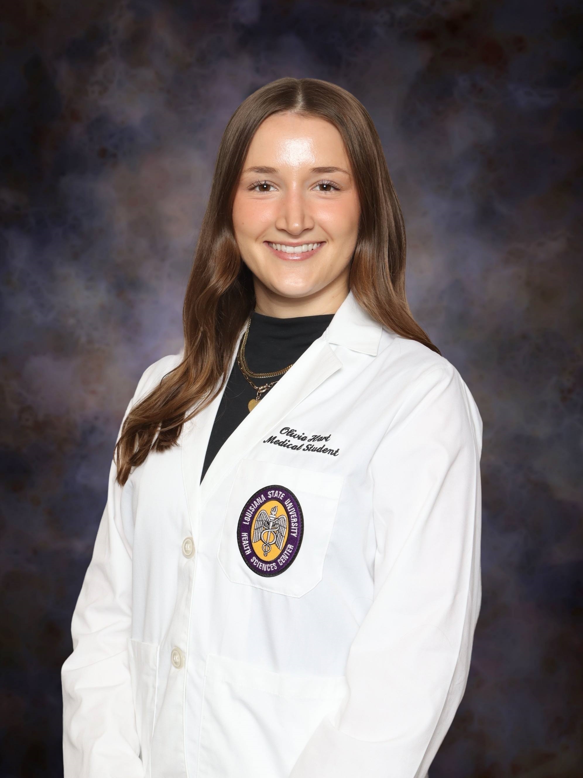 Head-and-shoulders studio portrait of a person with long hair wearing a white medical-style coat over a dark top, facing slightly toward the camera against a dark, mottled studio background. An institutional emblem and small embroidered text are visible on the coat.