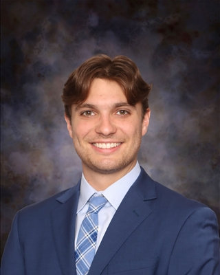A head-and-shoulders studio portrait of a person wearing a blue suit jacket, a light-colored dress shirt, and a patterned tie. The background is a softly blended dark studio backdrop with muted gray and brown tones, and the subject is facing forward in a formal, professional pose.
