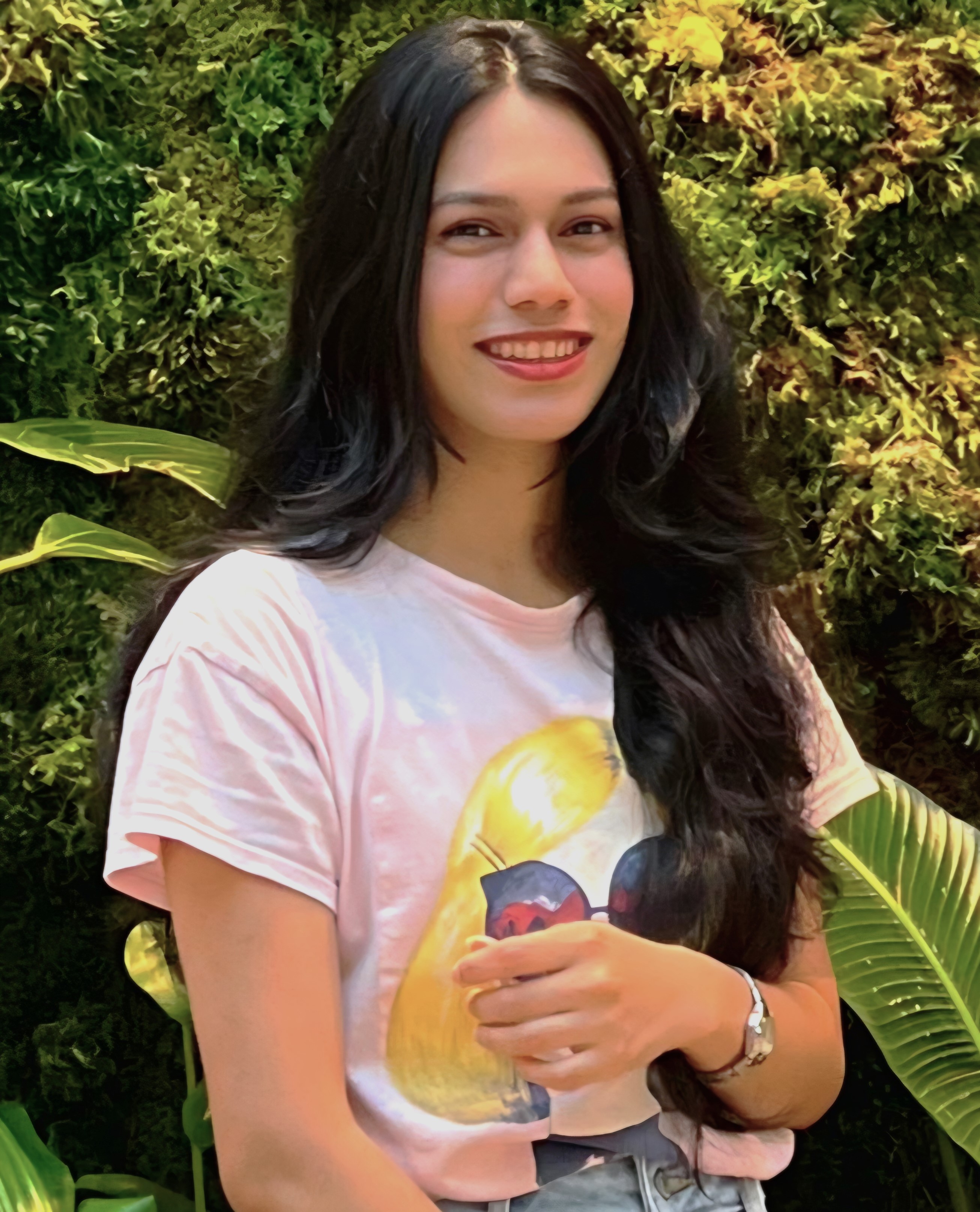 Outdoor portrait of a person with long dark hair wearing a light pink T‑shirt with a graphic on the front and light-colored jeans, standing in front of a dense green plant wall. 