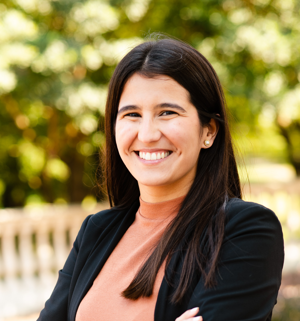Head-and-shoulders outdoor portrait of a person with long dark hair wearing a black blazer over a peach-colored top, standing with arms crossed. Sunlit trees and greenery appear softly blurred in the background.