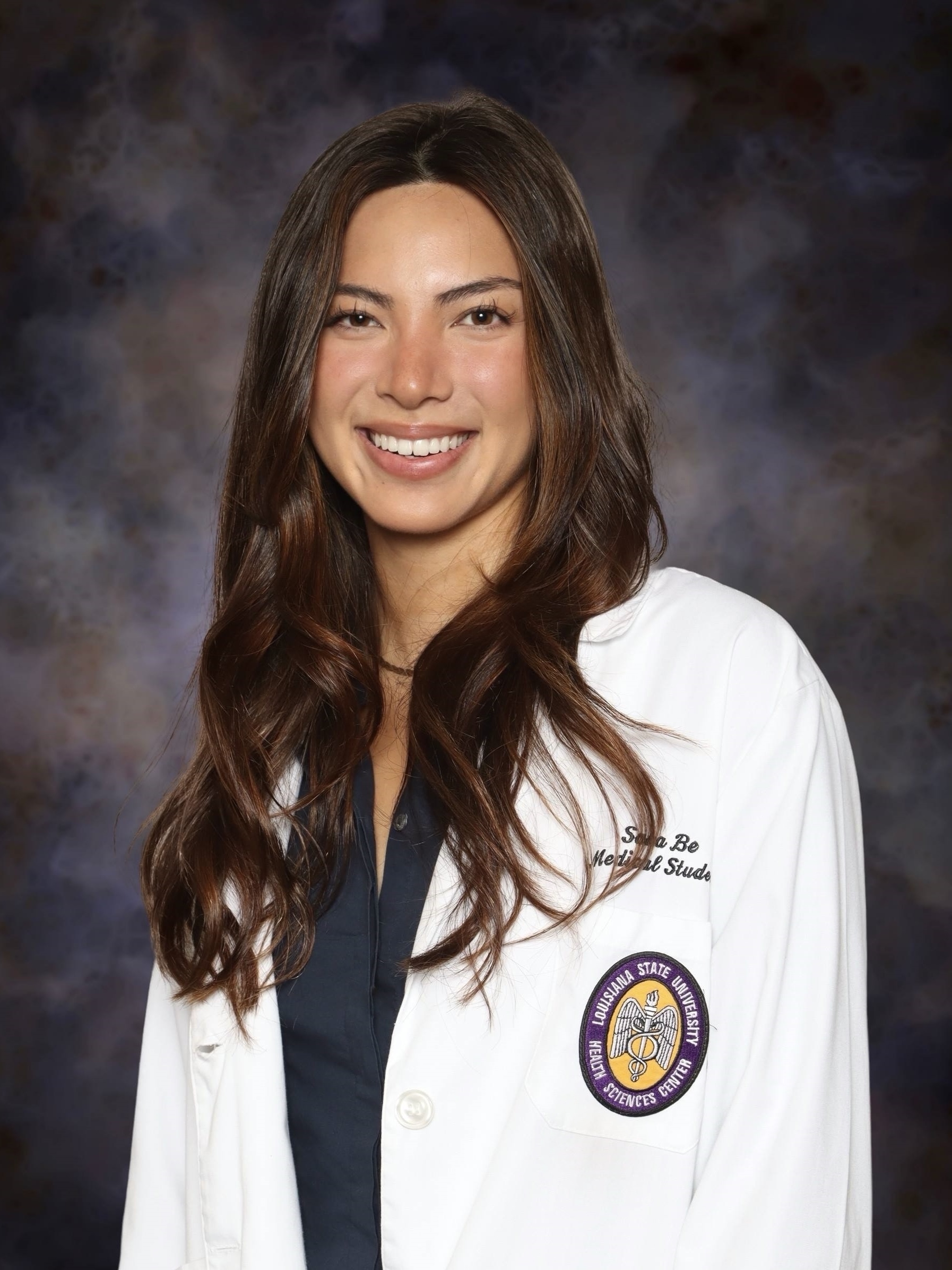 Head-and-shoulders studio portrait of a person with long wavy hair wearing a white medical-style coat over a dark top, facing slightly toward the camera against a dark, mottled studio background. An institutional emblem and small embroidered text are visible on the coat.