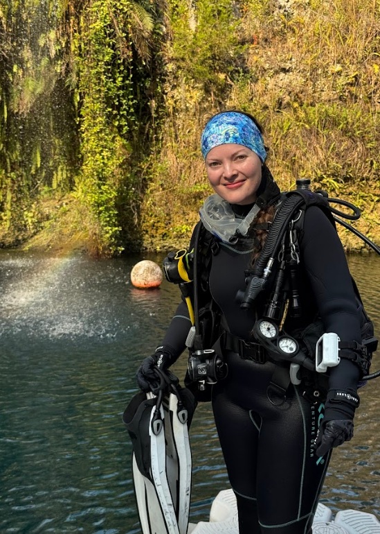 A person in full scuba diving gear stands on a dock at the edge of a clear freshwater spring, holding a pair of swim fins. The diver wears a black wetsuit, gloves, and a blue patterned head covering, with hoses, gauges, and tanks attached. Clear water is in the foreground, and a rocky, moss-covered wall with green vegetation rises behind the spring.