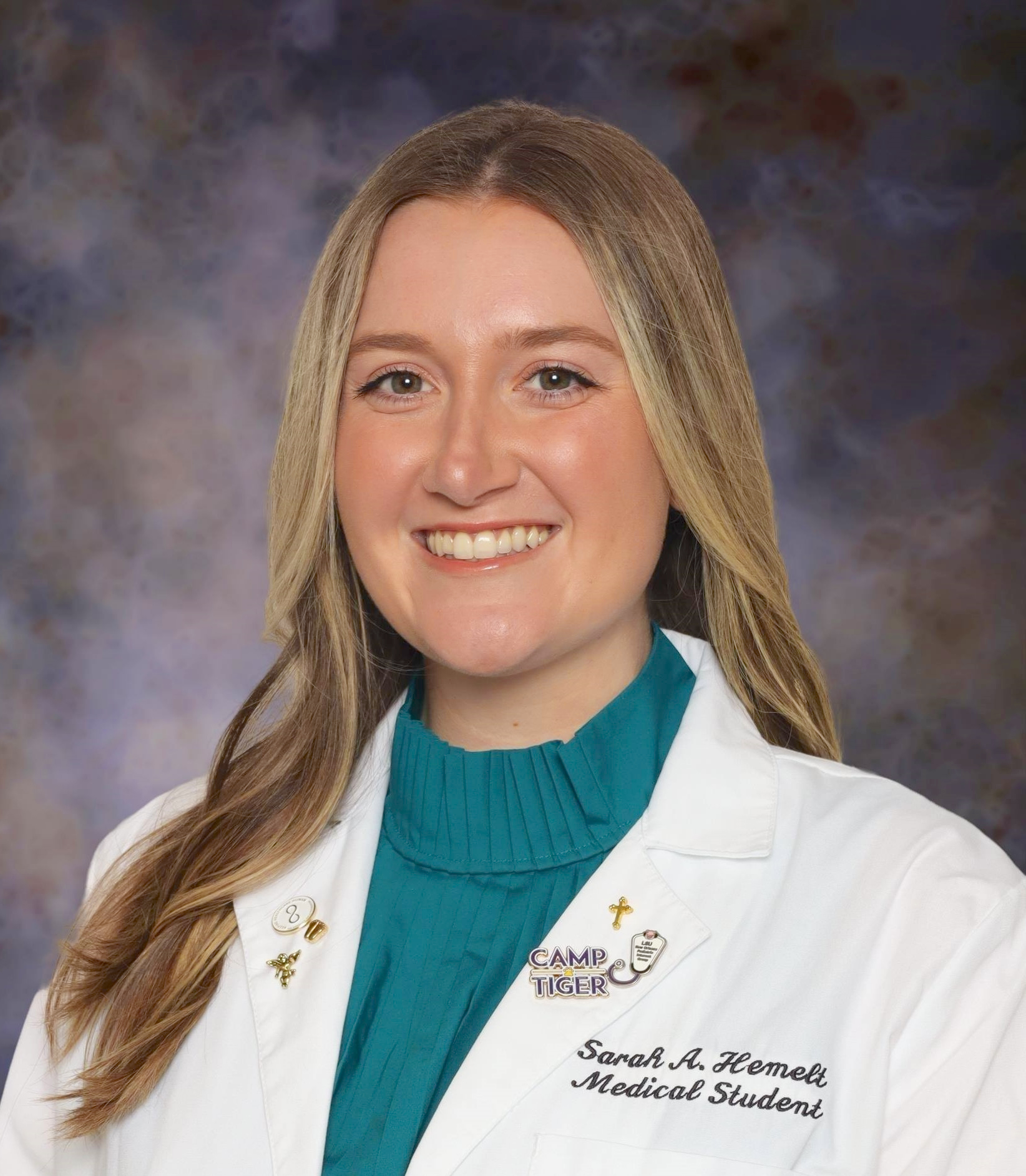 A professional studio portrait of a person wearing a white medical coat over a teal blouse. The medical coat has embroidered text and small pins on the lapel. The background is a softly blurred, mottled pattern in gray and purple tones, and the person is facing forward in a head-and-shoulders composition.