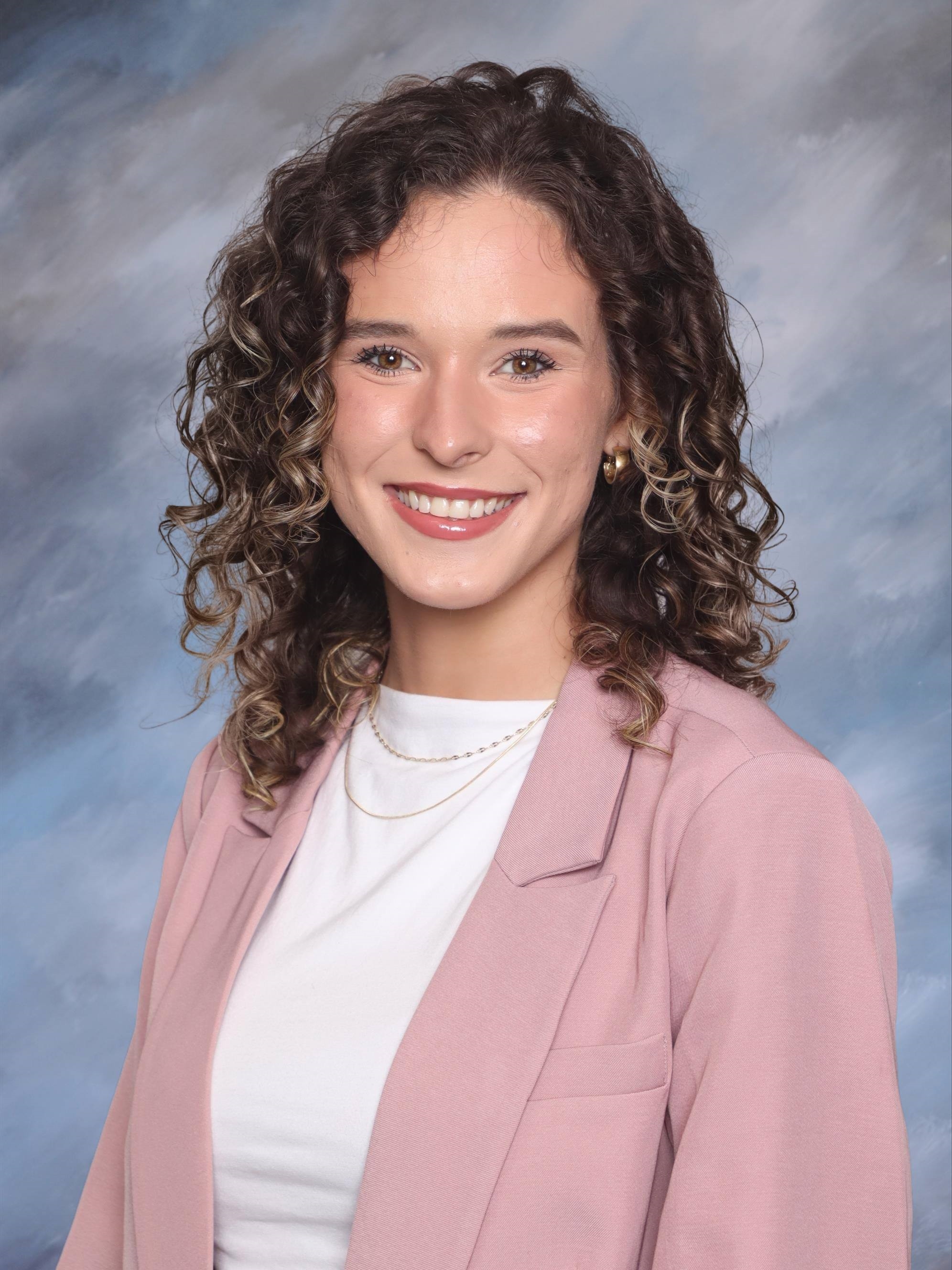 Head-and-shoulders studio portrait of a person with shoulder-length curly hair wearing a light pink blazer over a white top and layered necklaces, facing the camera against a softly blended blue and gray studio background.