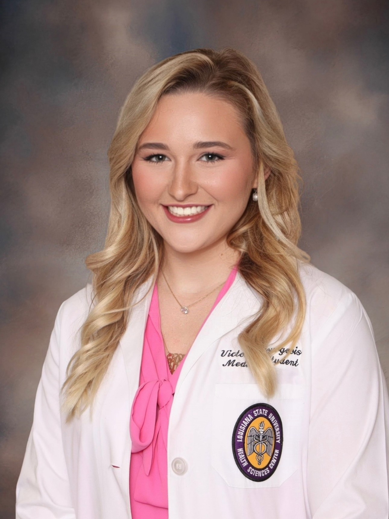 A head‑and‑shoulders studio portrait of a person wearing a white medical coat over a bright pink blouse with a tied neckline. The medical coat has embroidered text on the chest and a circular Louisiana State University Health Sciences Center patch on the lower front. The person is wearing a necklace and pearl earrings. The background is a softly blended studio backdrop in muted gray and brown tones, and the subject is facing forward in a professional pose.