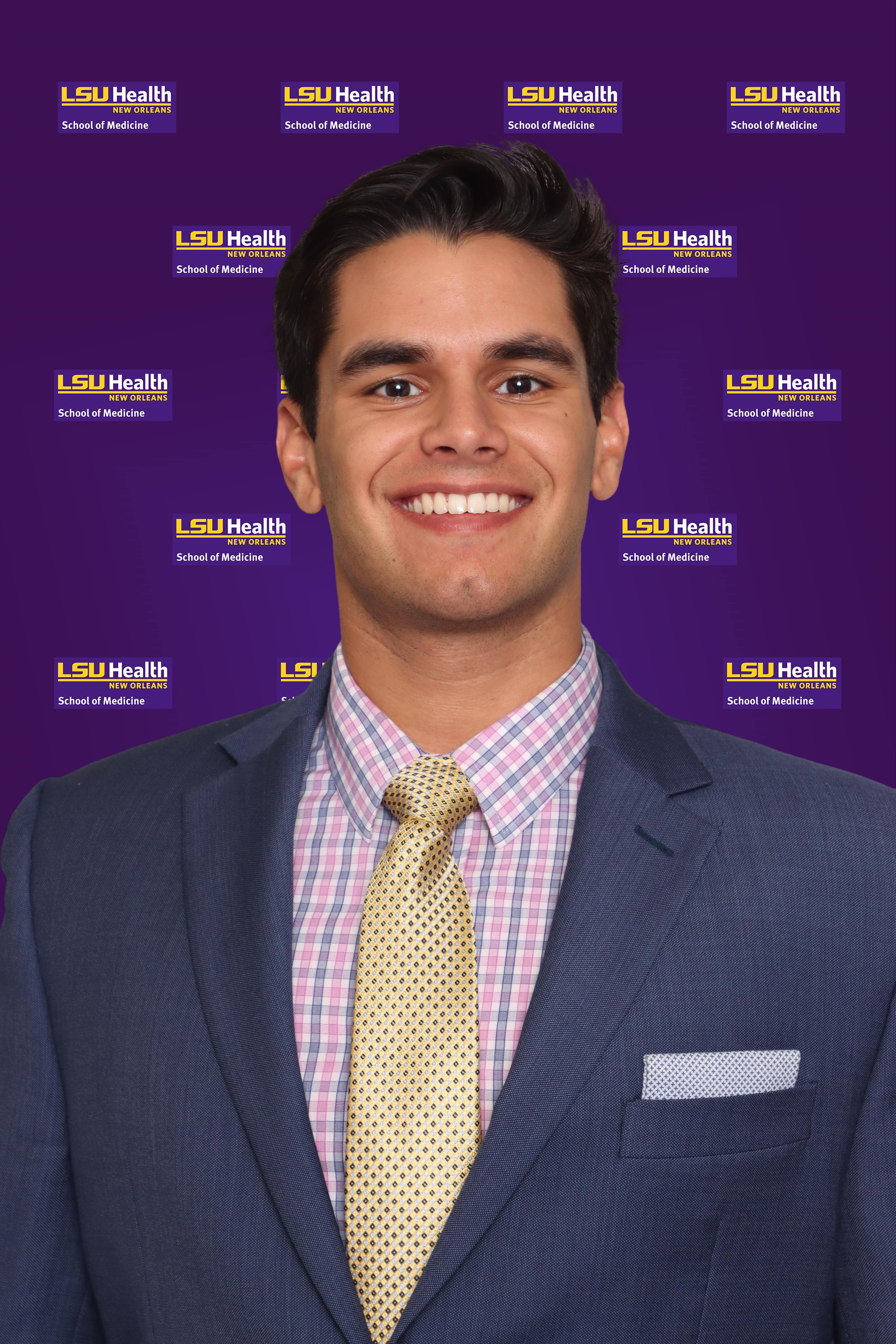 Head-and-shoulders studio portrait of a person wearing a textured blue suit jacket, patterned dress shirt, and light-colored tie, facing the camera against a purple backdrop repeating the text “LSU Health New Orleans – School of Medicine.”