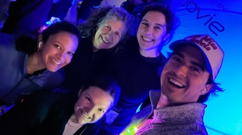 Five people gather for a selfie in a dimly lit indoor event space illuminated with blue and purple lighting. One person in the foreground holds the camera while the others lean in close to fit into the frame. Several attendees and tables are visible in the background. The scene has a casual, social atmosphere, with a glowing decorative item visible near the center and blue accent lighting throughout the room. Partial wall text is visible in the background but is not fully readable.