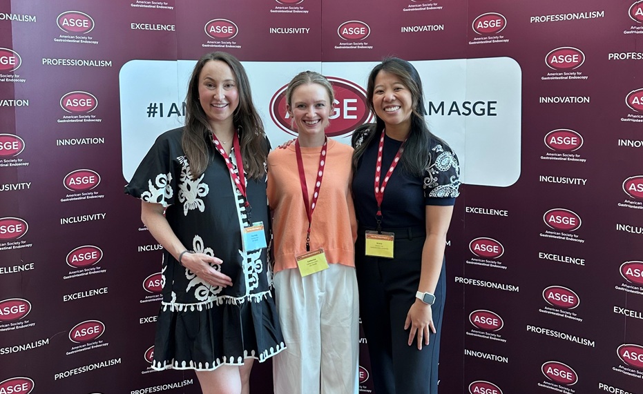 Three conference attendees wearing event badges and red lanyards pose in front of an ASGE (American Society for Gastrointestinal Endoscopy) step-and-repeat backdrop. The backdrop features repeated ASGE logos and the words “Professionalism,” “Excellence,” “Inclusivity,” and “Innovation,” along with the hashtag “#IAMASGE.” The attendees stand side by side facing the camera in a well-lit conference setting. One attendee wears a black-and-white patterned outfit, another wears a peach-colored top with light-colored pants, and the third wears a dark outfit with patterned sleeves and a smartwatch.