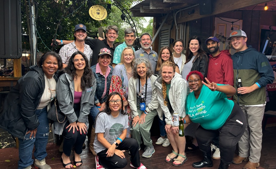 A group of approximately 15 people pose together on an outdoor wooden deck beneath trees and a covered patio area. The group is arranged in multiple rows, with some people standing and others crouching in front. Several attendees wear jackets, hats, and casual clothing, and one person holds a beverage bottle. The setting appears to be a social gathering at a restaurant or event venue, with wooden structures, string lights, and greenery visible in the background. A circular hanging sign and patio furnishings can be seen behind the group. Visible shirt text includes “NOLA Strong