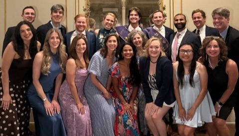 A group of 17 adults pose together indoors at a formal dinner or celebration. The group is arranged in two rows in front of an ornate gold-framed mirror and beige wall. Attendees wear semi-formal and formal attire, including suits, sport coats, dress shirts, cocktail dresses, and skirts in a variety of colors and patterns. Most are standing close together facing the camera, with some crouching or kneeling slightly in the front row. The room is warmly lit, creating a festive and celebratory atmosphere.