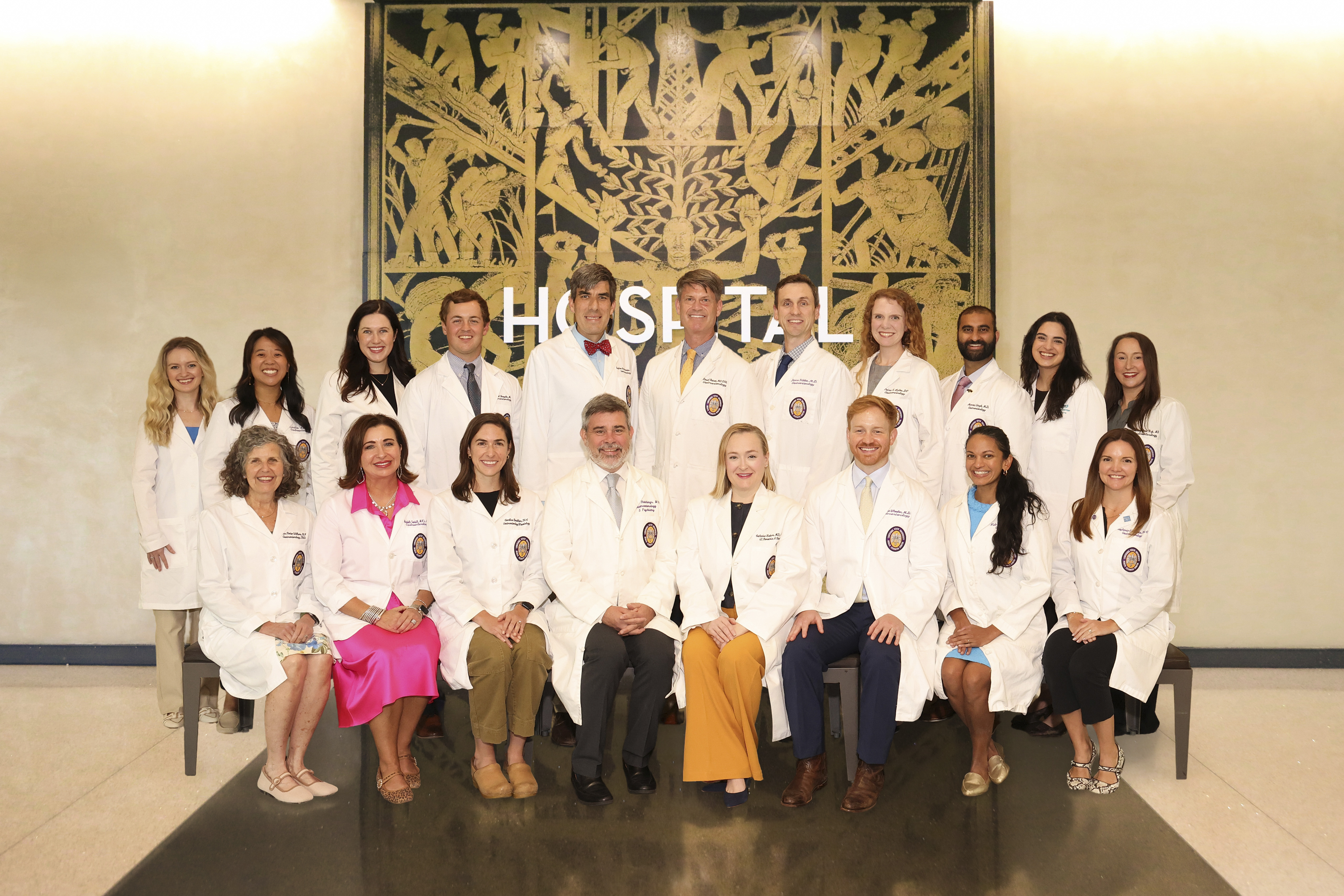 Group portrait of approximately 20 healthcare professionals wearing white lab coats, arranged in two rows in a hospital or academic medical center lobby. A large decorative wall panel with the word “HOSPITAL” is displayed behind the group.