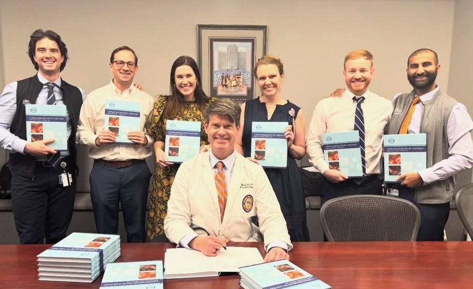 Seven people pose in a conference room around a large wooden table during a textbook release or signing event. Six individuals standing behind the table hold copies of a light blue medical textbook, while one individual seated at the center signs an open copy. Multiple stacks of the same textbook are arranged on the table in the foreground. A framed picture hangs on the wall behind the group, and office chairs surround the table. Visible text on the book cover includes Comprehensive Atlas of GI Endoscopy and Endoscopic Therapy, accompanied by several endoscopic images.