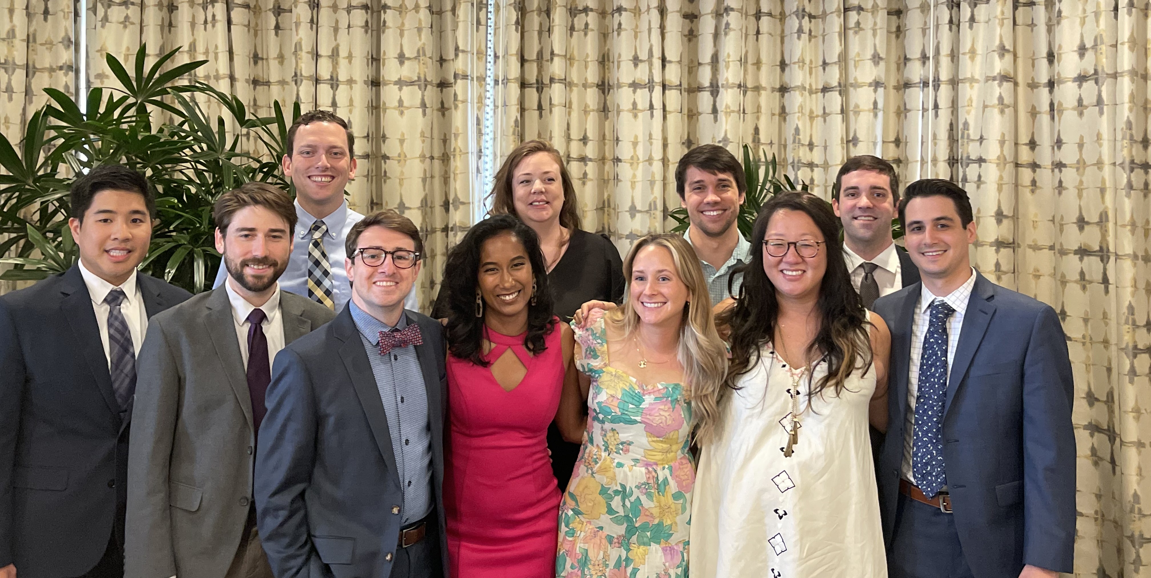 Group photo of eleven people dressed in business and semi-formal attire, posing indoors in front of patterned curtains and large potted plants.
