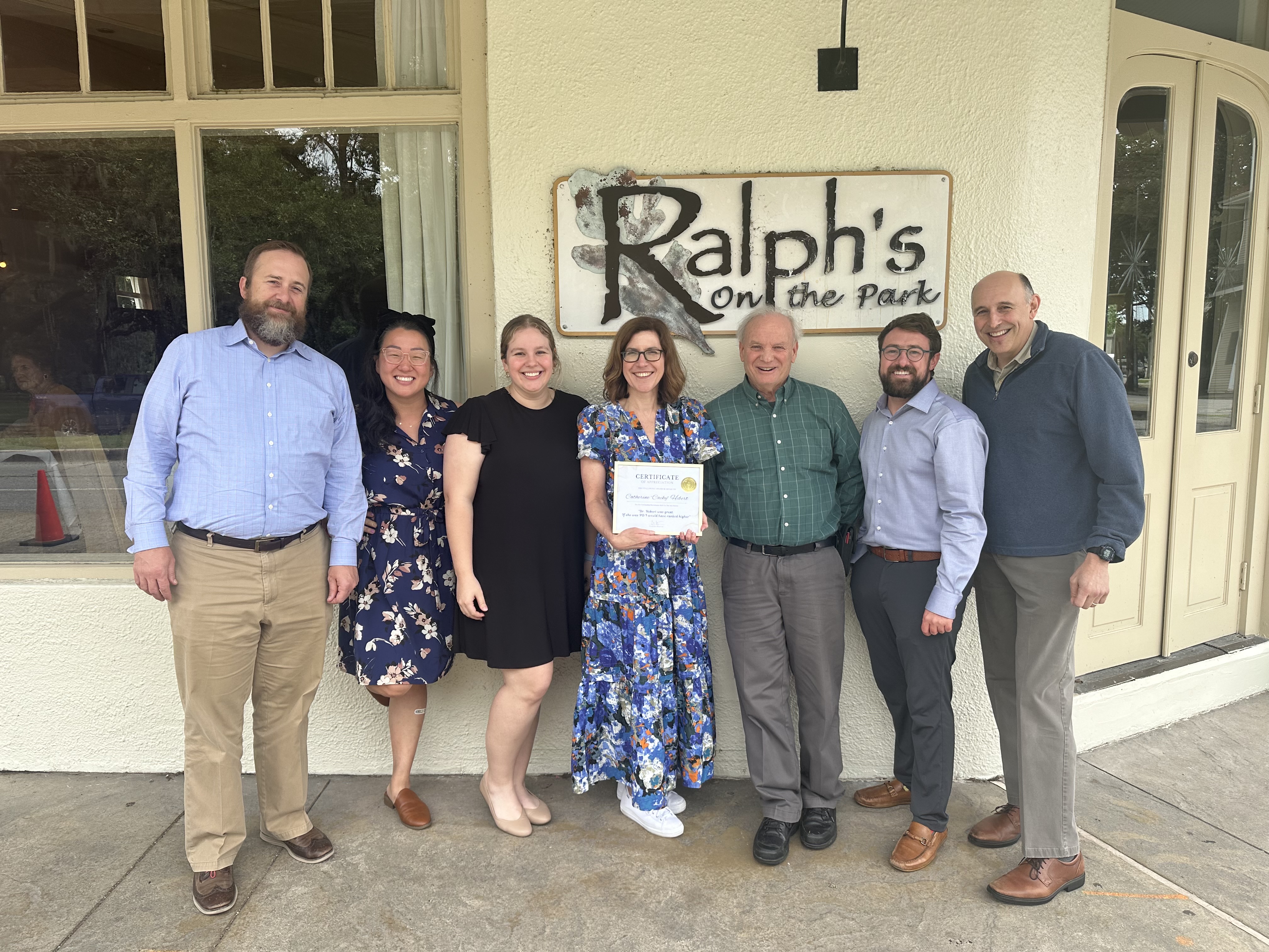 A group of seven people stand together outside in front of a restaurant sign that reads “Ralph’s on the Park.” One person in the center holds a certificate while the others stand close on either side. They are dressed in semi-formal or business-casual clothing, posing for a group photo on the sidewalk near the entrance.