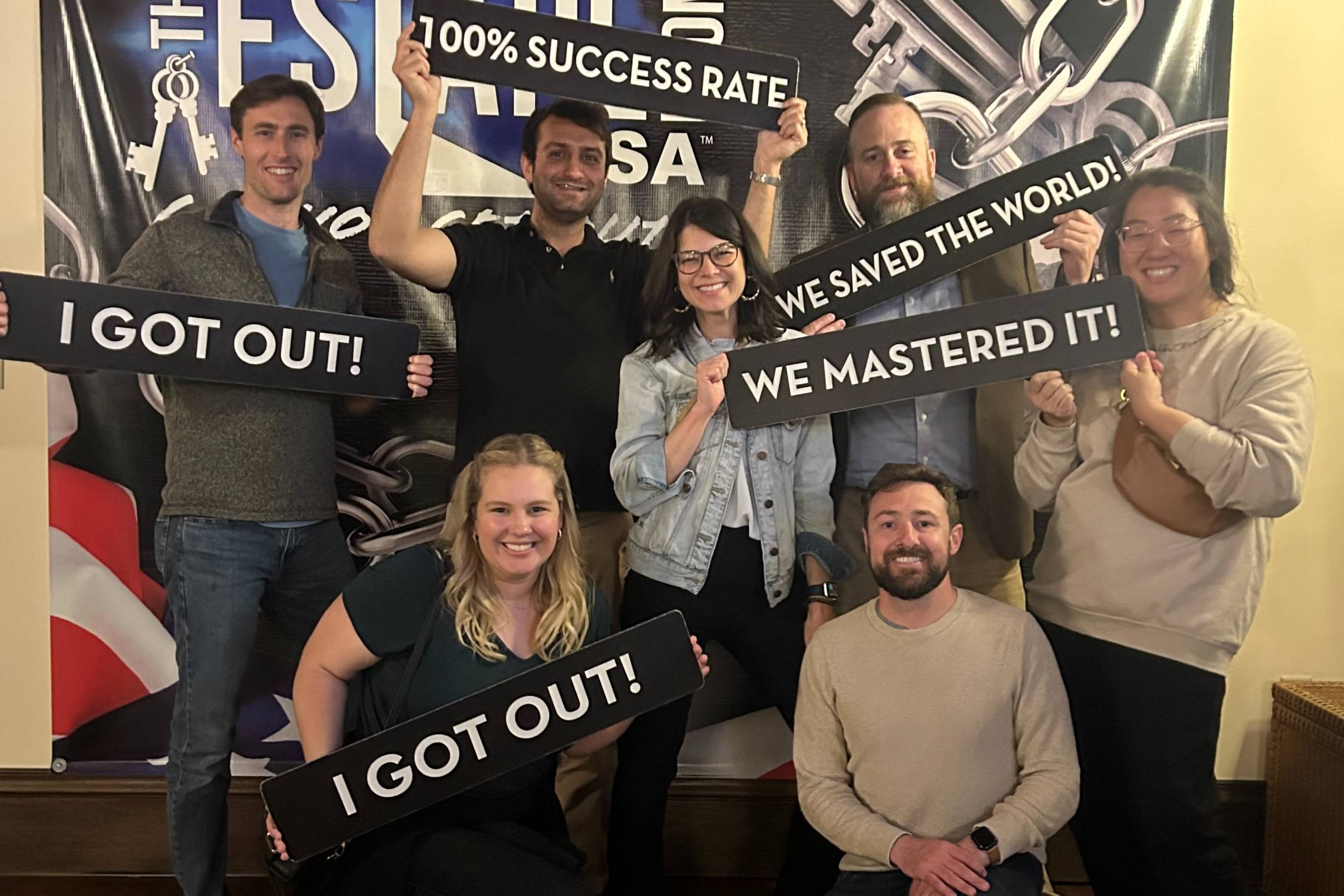 A group of people pose together in front of an escape‑room–themed backdrop. They hold signs with celebratory messages such as “I GOT OUT!,” “WE MASTERED IT!,” and “WE SAVED THE WORLD!” Some stand while others kneel in front, smiling and holding their signs proudly.
