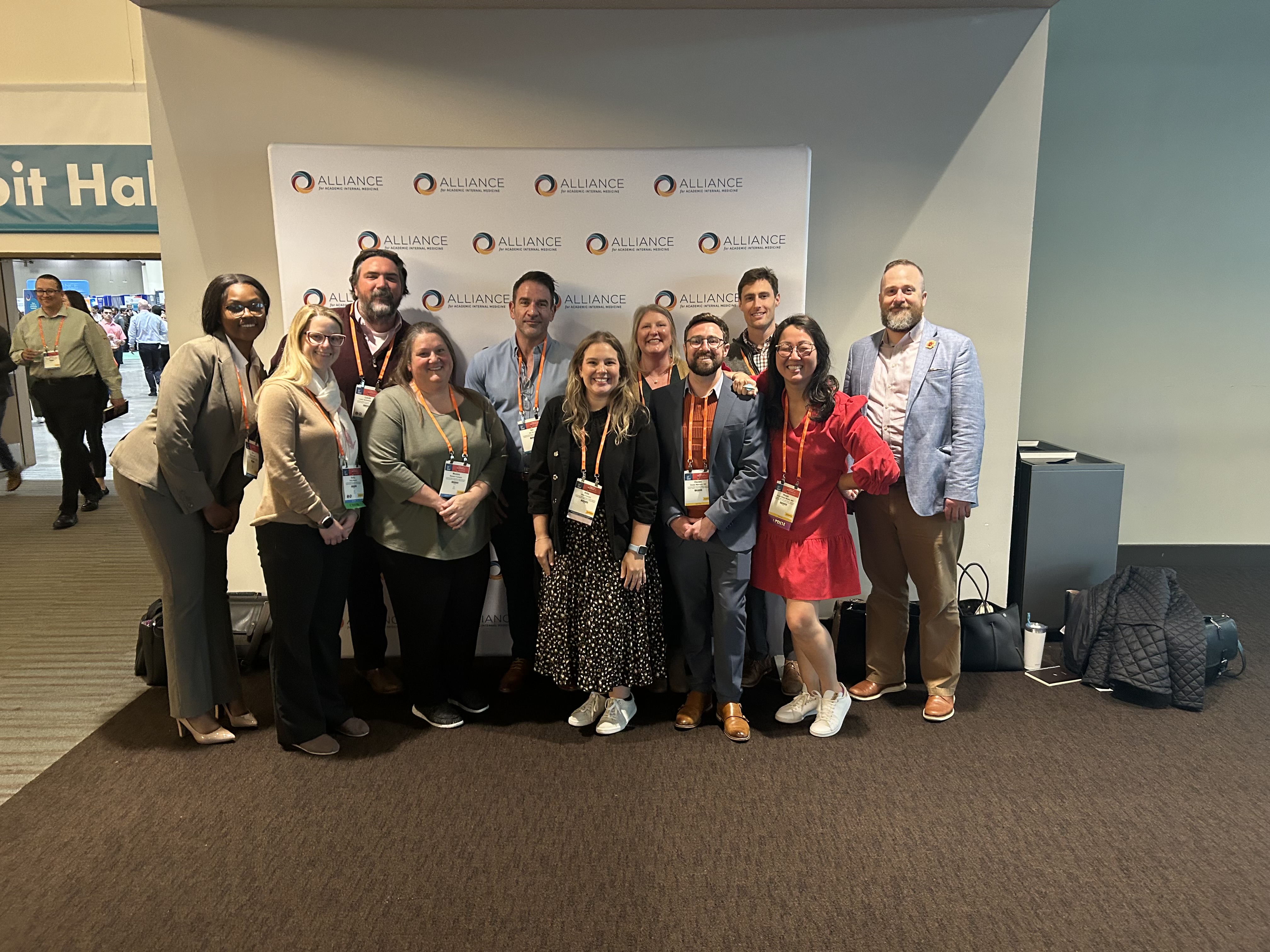 A group of people pose together in front of a white event backdrop printed with the Alliance logo. They are wearing business or business‑casual attire with conference lanyards and badges. The group stands closely in two rows, some kneeling in front, gathered for a group photo at a professional conference.