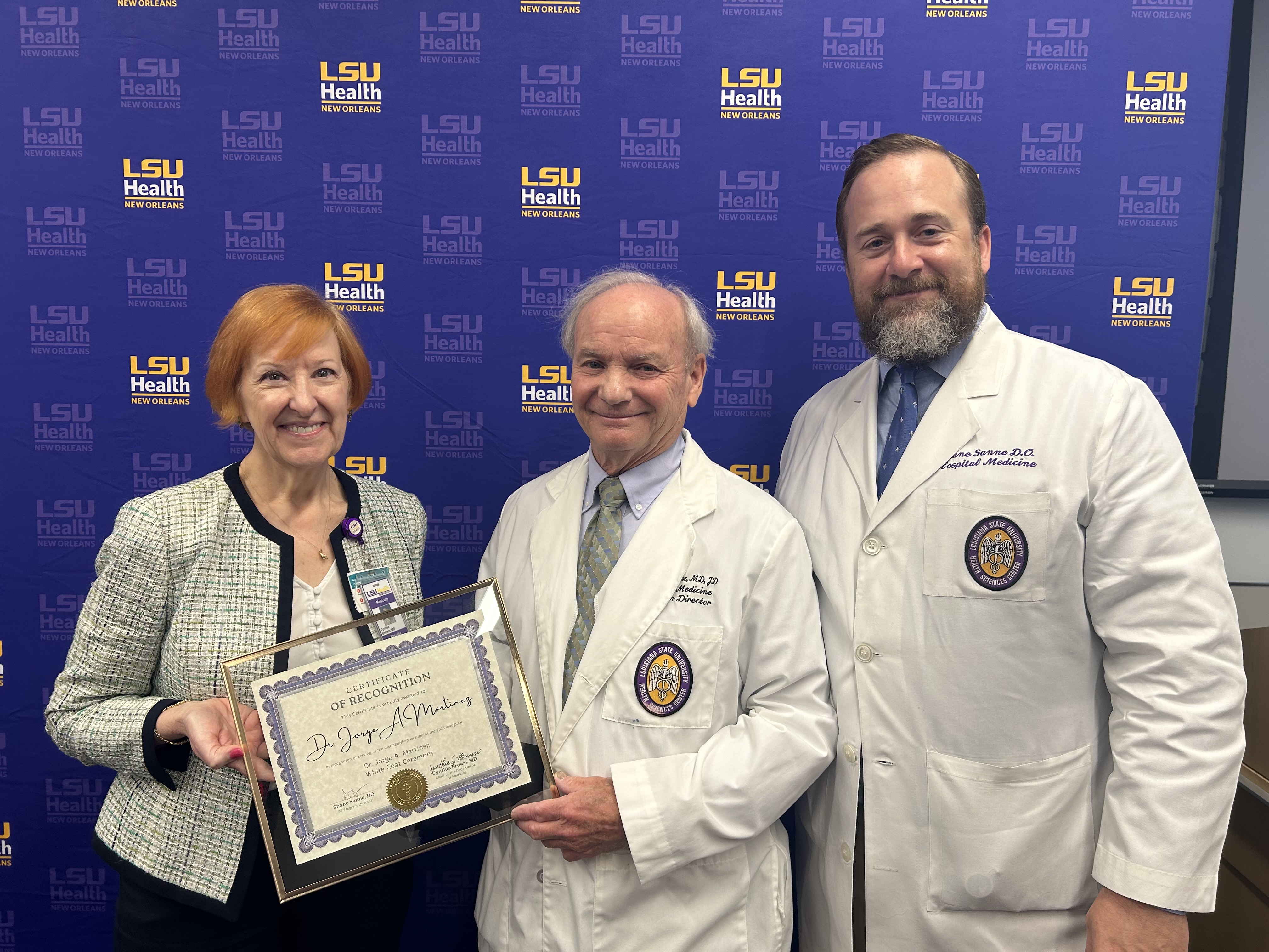 Three people stand together in front of a blue LSU Health backdrop. Two of them are wearing white medical coats, and one person is dressed in professional attire. The person in the center holds a framed certificate, and the group is posed closely for the photo.