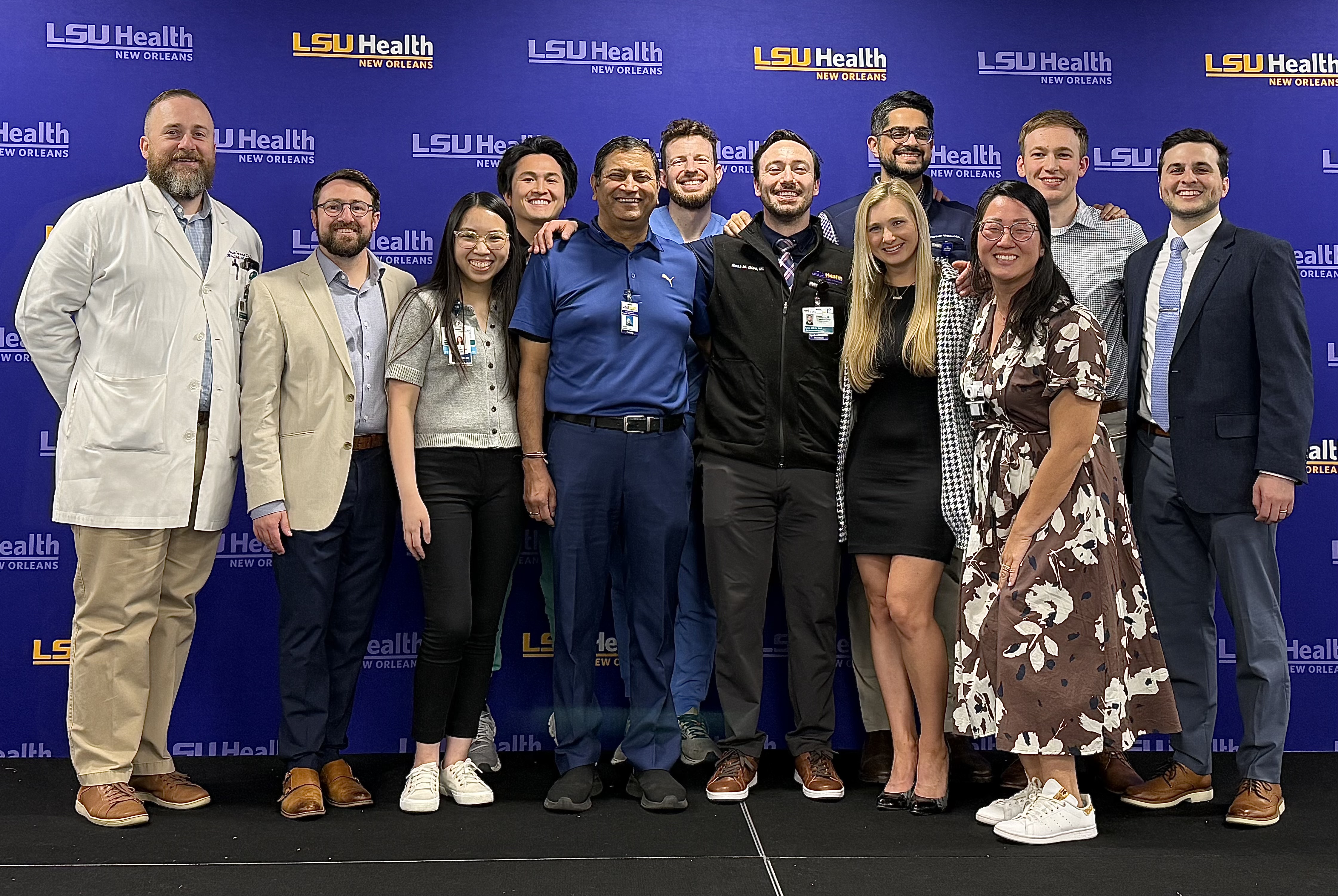 A group of people stand together in front of a blue LSU Health New Orleans backdrop. They are dressed in a mix of professional, business‑casual, and clinical attire, including suits, dresses, scrubs, and a white coat. The group poses closely for a group photo on a stage-like platform.