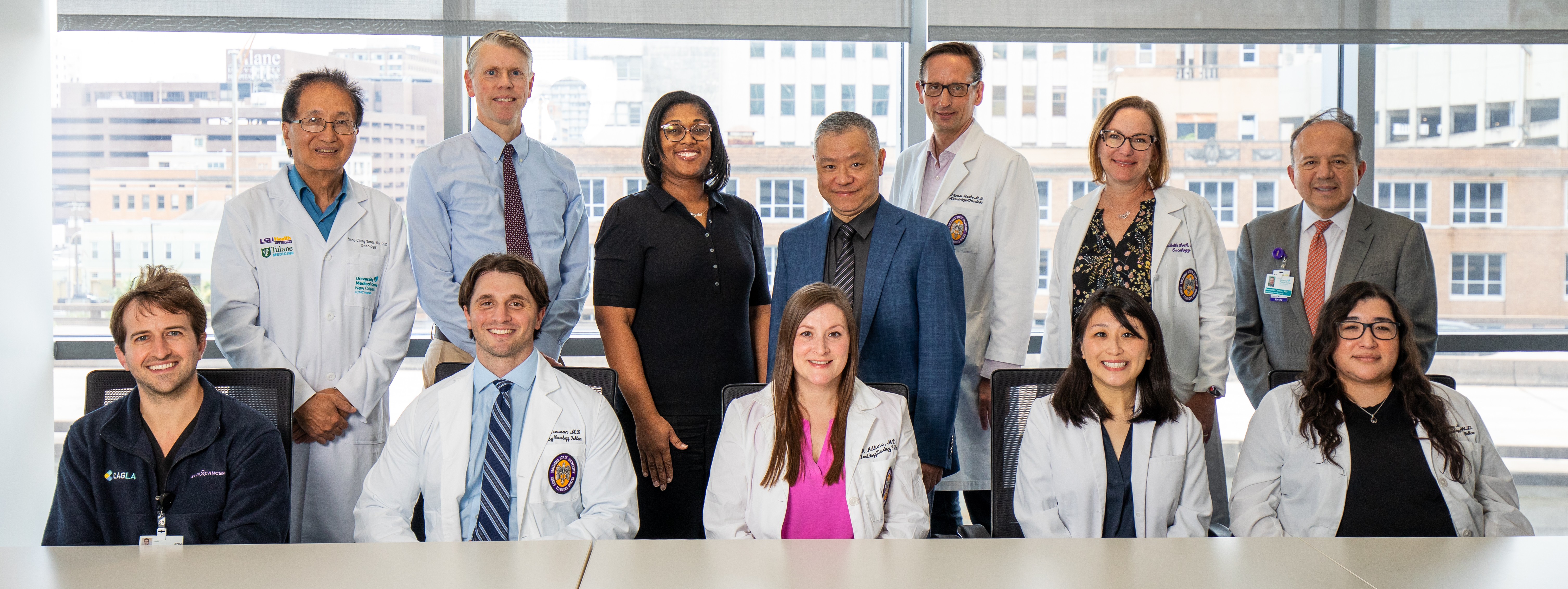 Group portrait of 11 healthcare and research professionals in a conference room. Five people are seated in the front row and six are standing behind them. Several individuals wear white medical lab coats, while others wear business or professional attire. Large floor-to-ceiling windows behind the group reveal a cityscape with office buildings. The group faces the camera around a conference table in a bright, well-lit office setting.