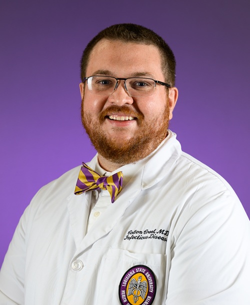 Professional studio portrait of a person wearing eyeglasses, a white medical coat, and a purple-and-gold striped bow tie, posed against a solid purple background. The white coat features an embroidered name and specialty on the chest and a Louisiana State University Health Sciences Center emblem on the lower pocket. The person is facing the camera in a three-quarter pose with head and shoulders visible.