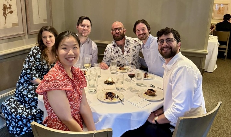 Six people seated around a dining table sharing a meal in a restaurant or banquet setting.