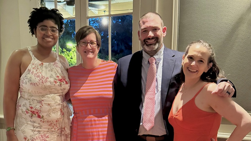 Four people pose together at an indoor social event in front of a window, with one person in a suit and tie and others in dresses.