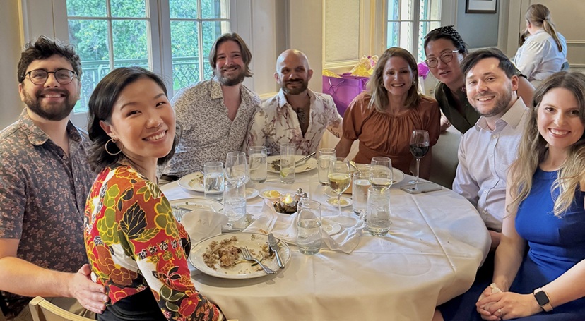 Eight people gathered around a dining table in a banquet room, posing for a group photo with food and drinks on the table.