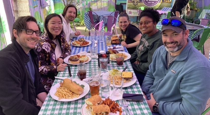 Six people share a meal at an outdoor restaurant table with sandwiches, fries, and drinks on a colorful patio.