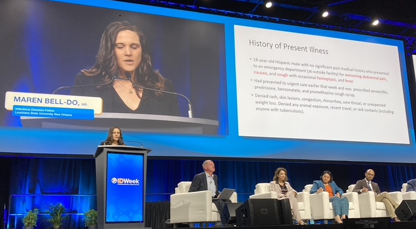 Speaker presents a patient case study at a medical conference while a panel of experts sits onstage and slides are displayed on large screens.