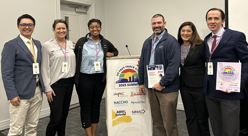 Six attendees pose beside a podium at the US Fast-Track Cities 2025 Summit, displaying conference badges and event materials.