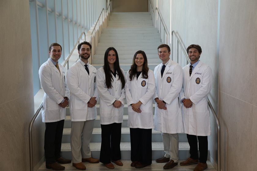 Six people wearing white medical lab coats stand together on an indoor staircase in a modern building. The group is arranged in a single row at the base of the stairs, with professional attire visible beneath the coats. Tall windows along the left side provide natural light, and metal handrails line both sides of the staircase. The white stairway extends upward behind the group, creating a symmetrical and professional backdrop.