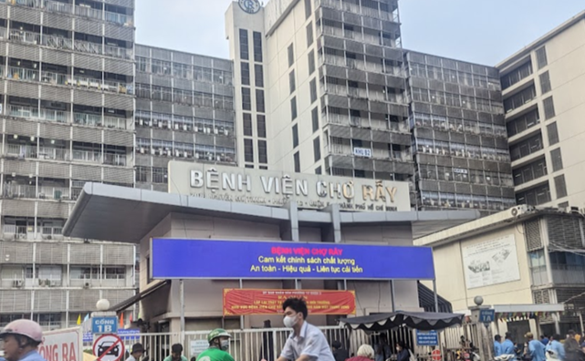 A large multi‑story hospital building with the sign ‘Bệnh Viện Chợ Rẫy’ above the entrance. A wide electronic display board and red banner are positioned at the front of the building. Numerous motorbikes and scooters are parked and moving in the foreground, with several people riding or walking near the hospital entrance. The setting appears busy, with surrounding buildings and street activity visible.