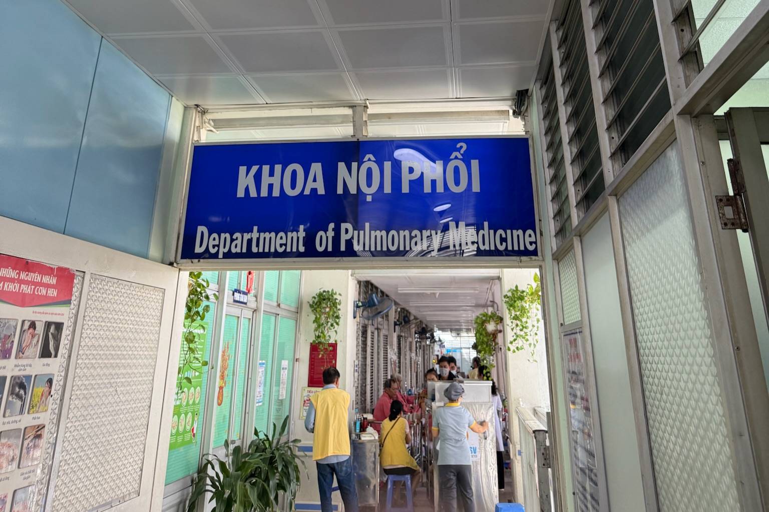 A corridor in a medical facility with a large blue sign overhead that reads ‘Khoa Nội Phổi’ and ‘Department of Pulmonary Medicine.’ Below the sign, several people are seated and standing along the hallway. The corridor has potted plants hanging on the walls, light-colored flooring, and glass-panel doors and windows lining both sides.