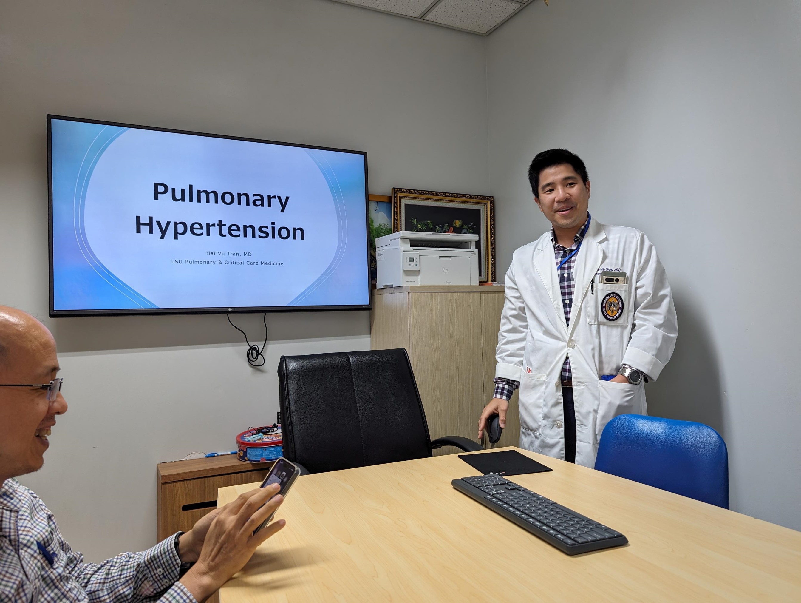 A person in a white medical coat stands at the front of a small meeting room next to a wall-mounted screen displaying the title ‘Pulmonary Hypertension.’ Another person sits at the table in the foreground, holding a smartphone. The table has a keyboard and mouse on it, and a printer and framed pictures sit on a cabinet behind the standing person. The room has plain walls and office-style lighting.