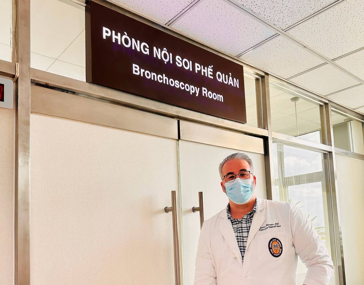 A person wearing a white medical coat and a face mask stands in front of a set of light‑colored double doors. Above the doors is a sign with Vietnamese text reading ‘Phòng Nội Soi Phế Quản’ and English text reading ‘Bronchoscopy Room.’ The setting appears to be a hospital hallway with a drop ceiling and fluorescent lighting.