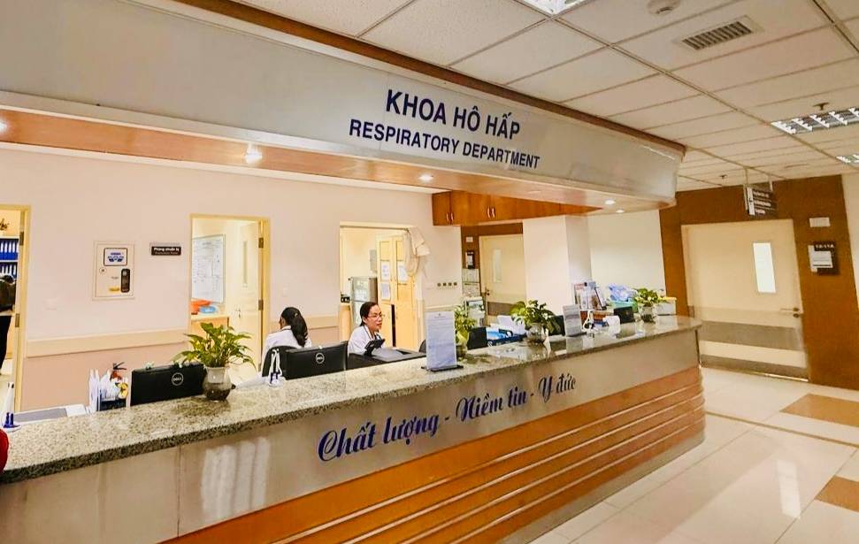 A hospital reception area with a long counter labeled ‘Khoa Hô Hấp’ and ‘Respiratory Department’ on the overhead sign. Several staff members sit behind the counter working at computers. Potted plants are placed along the counter, and informational papers and office supplies are visible. The surrounding hallway has tiled floors, overhead lighting, and multiple doorways leading to other rooms.
