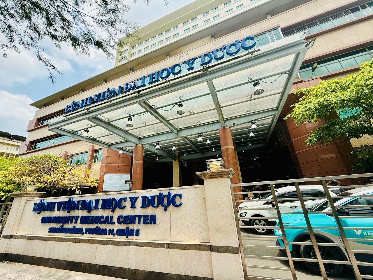 A large multi‑story hospital building with a glass canopy over the main entrance. Blue signage on the building reads ‘Bệnh Viện Đại Học Y Dược’ and the English translation ‘University Medical Center’ is displayed on the front wall along with the address. Several vehicles, including a turquoise taxi, are parked or driving in front of the building. Trees frame the scene, and the sky is partly cloudy.