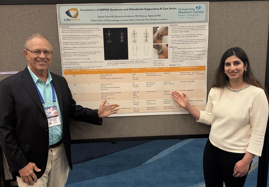 Two conference attendees stand beside a research poster during a poster session, each gesturing toward the display. The poster is mounted on a presentation board and includes text, images, and a data table.