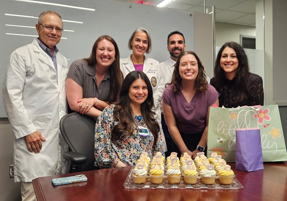 Group photo taken in an office conference room showing seven people gathered around a table. One person is seated in an office chair while six others stand behind and beside the chair. A tray of decorated cupcakes and gift bags are placed on the table in the foreground, suggesting a celebration or special occasion. Two people are wearing white lab coats, and a large whiteboard is visible in the background. A smartphone rests on the table near the seated person.