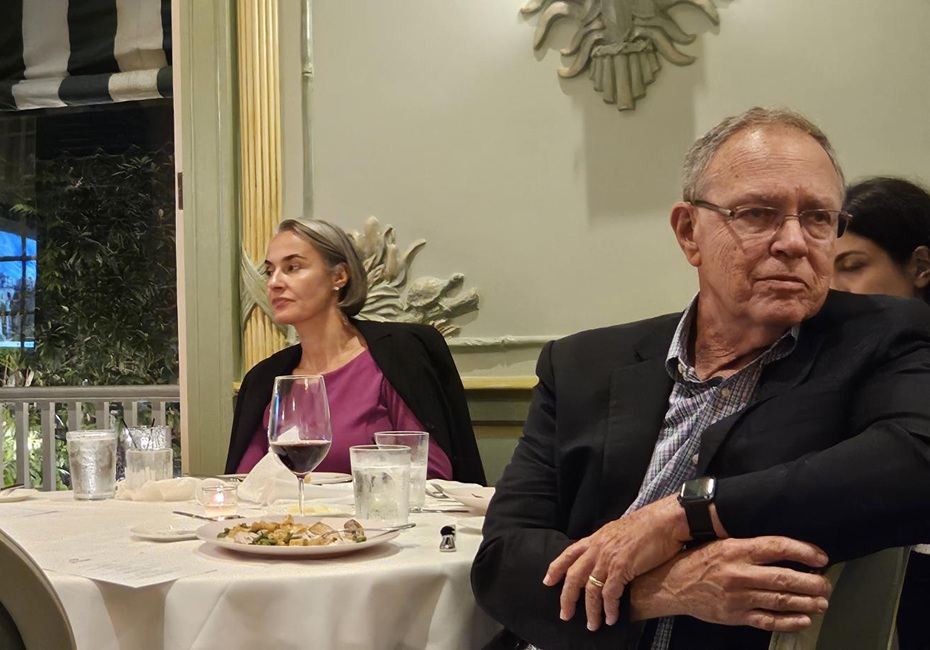 Attendees are seated at a round banquet table during a meeting or dinner event. In the foreground, a person wearing a dark jacket and glasses sits with arms folded, facing toward the right side of the room. Another attendee in a purple top and dark blazer sits across the table near a window. The table is set with water glasses, a glass of red wine, plates, and dining utensils. Decorative wall accents, soft lighting, and a window overlooking an outdoor area create a formal dining atmosphere.