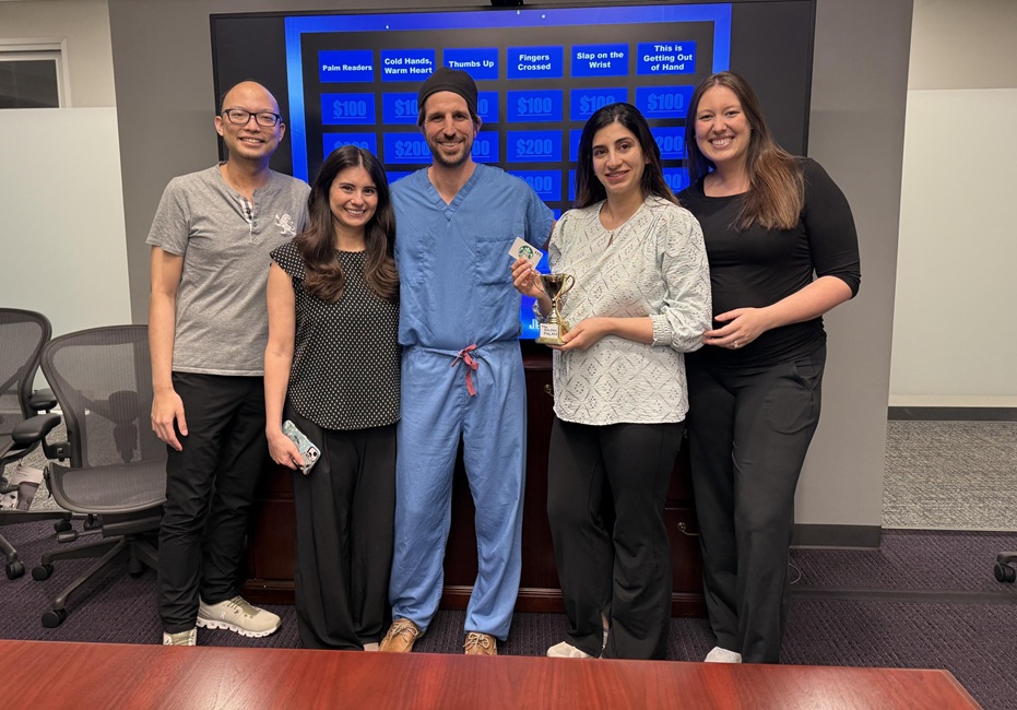 Five people pose together in a conference room, with one person holding a small trophy. A large screen behind the group displays a Jeopardy-style game board used for an educational activity.