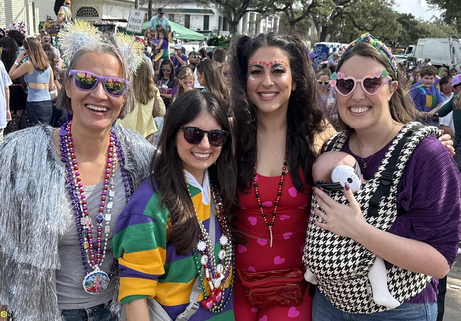 Four people wearing colorful outfits, beads, and festive accessories pose together at a Mardi Gras parade. One person is carrying an infant in a front baby carrier.