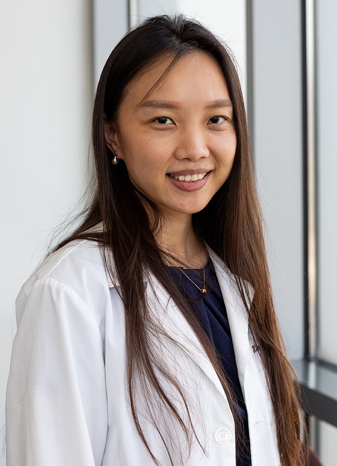 Professional head-and-shoulders portrait of a person with long dark hair wearing a white lab coat over a dark top. The person is standing indoors near large windows, facing the camera. Natural light illuminates the scene, and the background features a bright, modern interior with glass panels and vertical window frames.